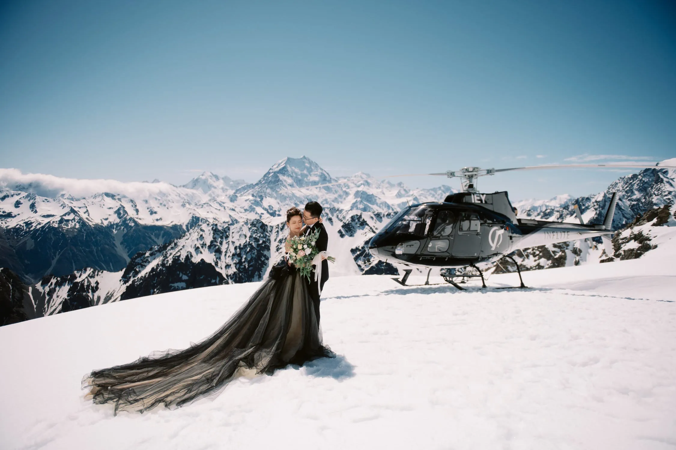 Queenstown Tekapo New Zealand Heli Wedding Elopement Pre-Wedding Shoot Photographer クイーンズタウン　テカポ　ニュージーランド　エロープメント 前撮り　フォトウェディング　結婚式 | A couple embracing on snow-covered terrain with a helicopter in the background at Tekapo, New Zealand, suggesting a pre-wedding shoot or alpine photoshoot.